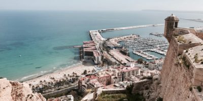 A sandy beach lined with palm trees and a busy marina, seen from a stone fortress wall high above a seaside city