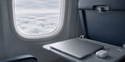 An airplane window over a white cloud layer, with a closed laptop and earbuds case on the tray table