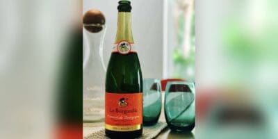 The La Burgondie Crémant de Bourgogne bottle, green glass with an orange label, shown whole against a blurred version of the same photograph